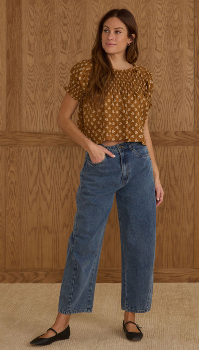Woman wearing a patterned top and blue jeans against a wooden paneled wall. Rylee + Cru Barrel Pant — Indigo Denim