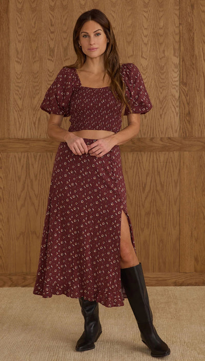 Woman wearing a burgundy patterned dress with a slit and black knee-high boots against a wooden paneled wall.