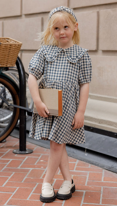 Young girl in a checkered dress holding a book on a sidewalk. Camille Dress | Navy Check | Rylee and Cru
