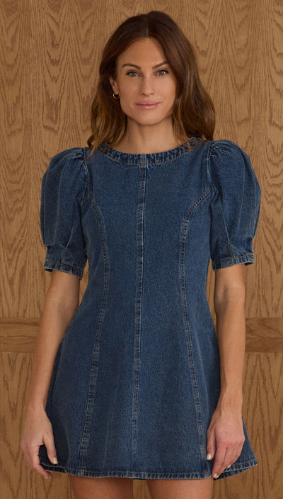 Woman wearing a denim dress with puff sleeves against a wooden paneled wall. Levi denim dress by Rylee and Cru.