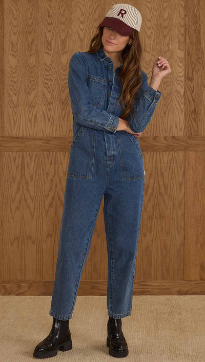 Woman wearing a blue denim jumpsuit and a cap with a logo against a wooden paneled wall. Rylee + Cru Long Sleeve Coverall – Indigo Denim