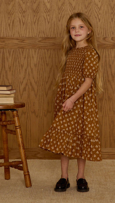 Young girl in a patterned dress standing in front of a wooden paneled wall. Lydia Dress | Golden Fleur | Rylee and Cru