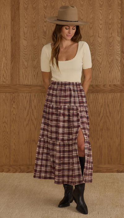 Woman wearing a plaid skirt, white top, and brown hat against a wooden paneled wall. Tiered Midi Skirt – Burgundy Plaid | Rylee + Cru